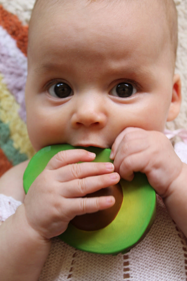 A baby holding an Oli & Carol Arnold avocado teething toy made of all-natural rubber