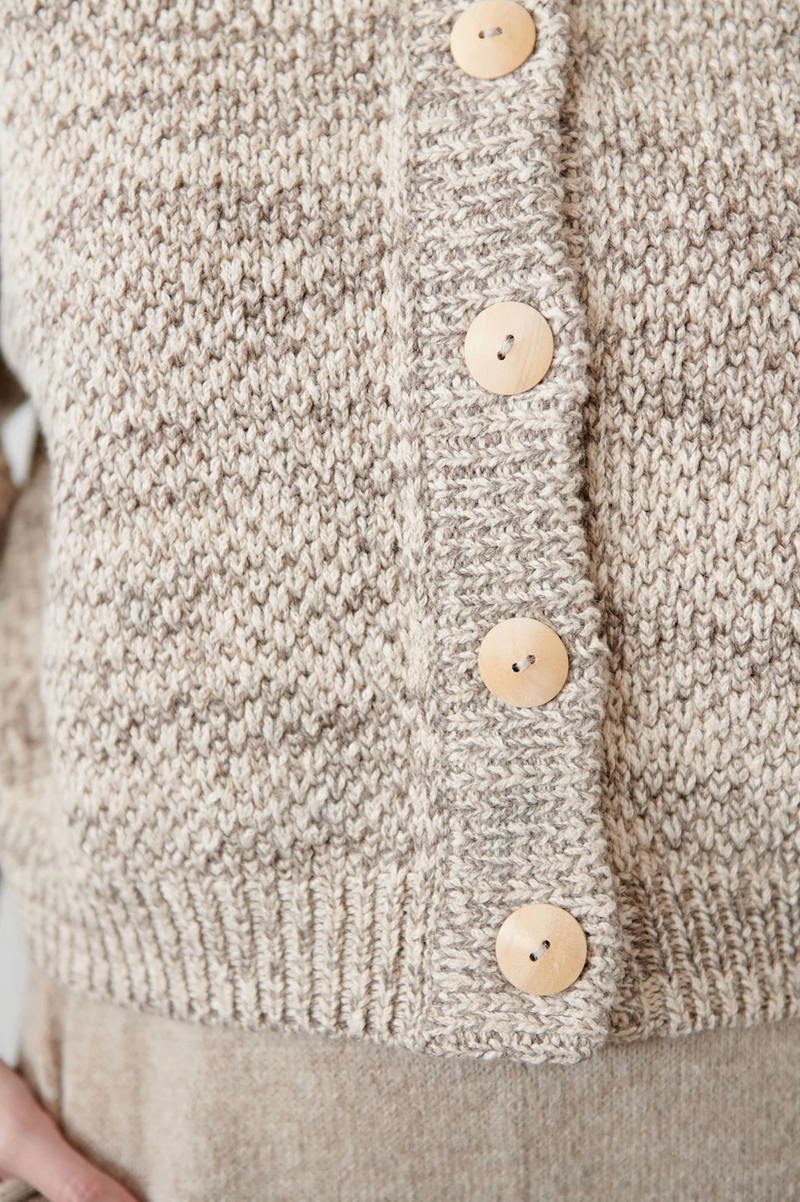 A close up view of a model wearing a cropped wide cardigan featuring unvarnished wood buttons and a high, rounded collar made of wool/alpaca blend chain yarn