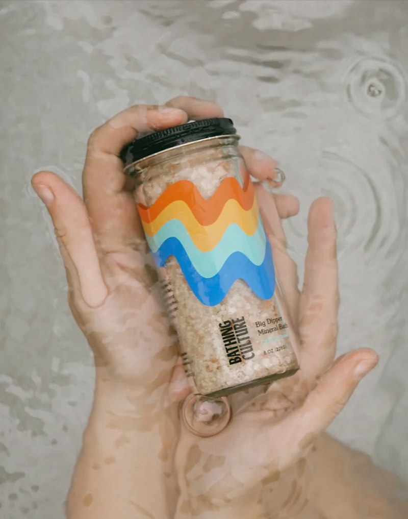 A person holding a container of Bathing Culture Big Dipper Mineral Bath, made from a blend of mineral-rich salts, green clay, and jojoba oil