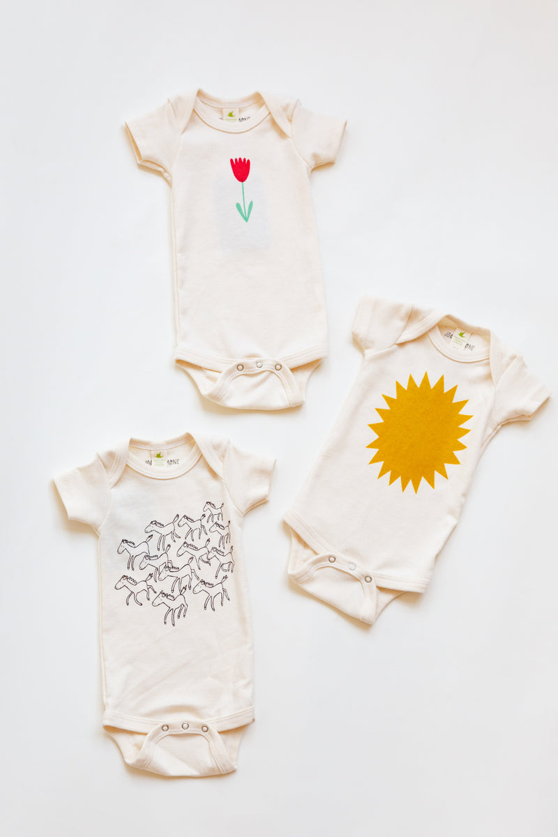 A Joan Ramone baby sunshine onesie printed on 100% organic cotton with non-toxic, water-based ink, laying flat on a table with a couple other onesies
