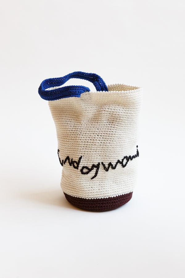 A hand-crocheted bucket bag with Sunday Morning embroidered letters on display