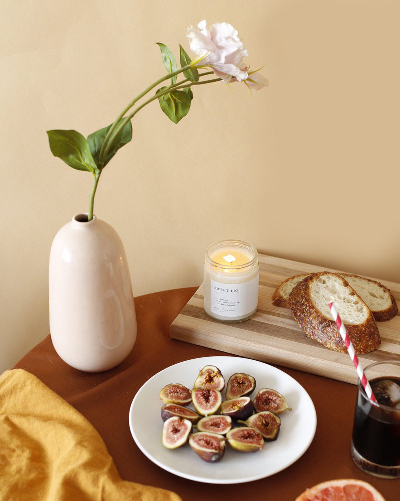 A Brooklyn Candle Studio jar in Sweet Fig scent on a table with bread, figs, and flowers